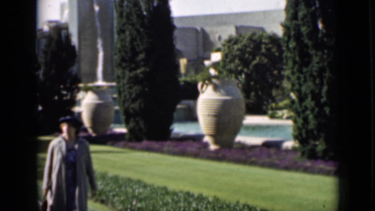 Woman in a hat walking on lawn beside trees, urns, and a shallow pool.
