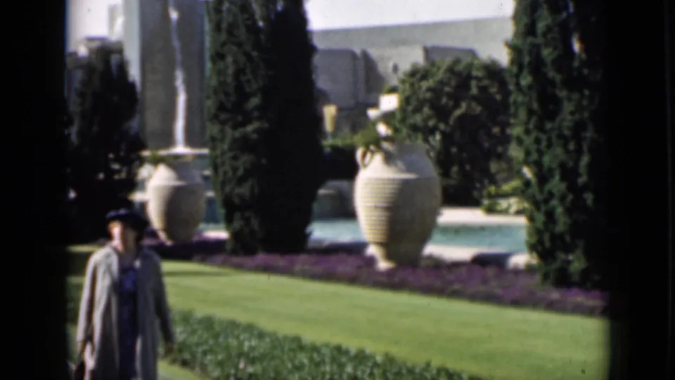 1937 San Francisco frame of a woman walking through formal garden grounds with tall trees, large urns, and a pool edge.
