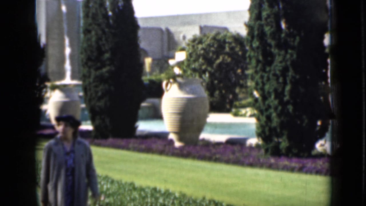 Home-movie view of a woman walking across landscaped San Francisco grounds with urns, cypress-like trees, and planting beds.