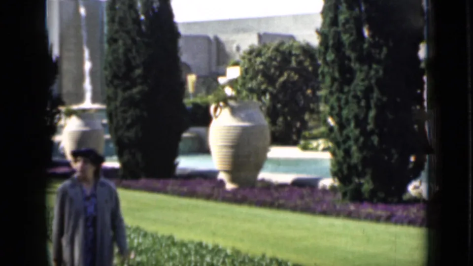 Home-movie view of a woman walking across landscaped San Francisco grounds with urns, cypress-like trees, and planting beds.