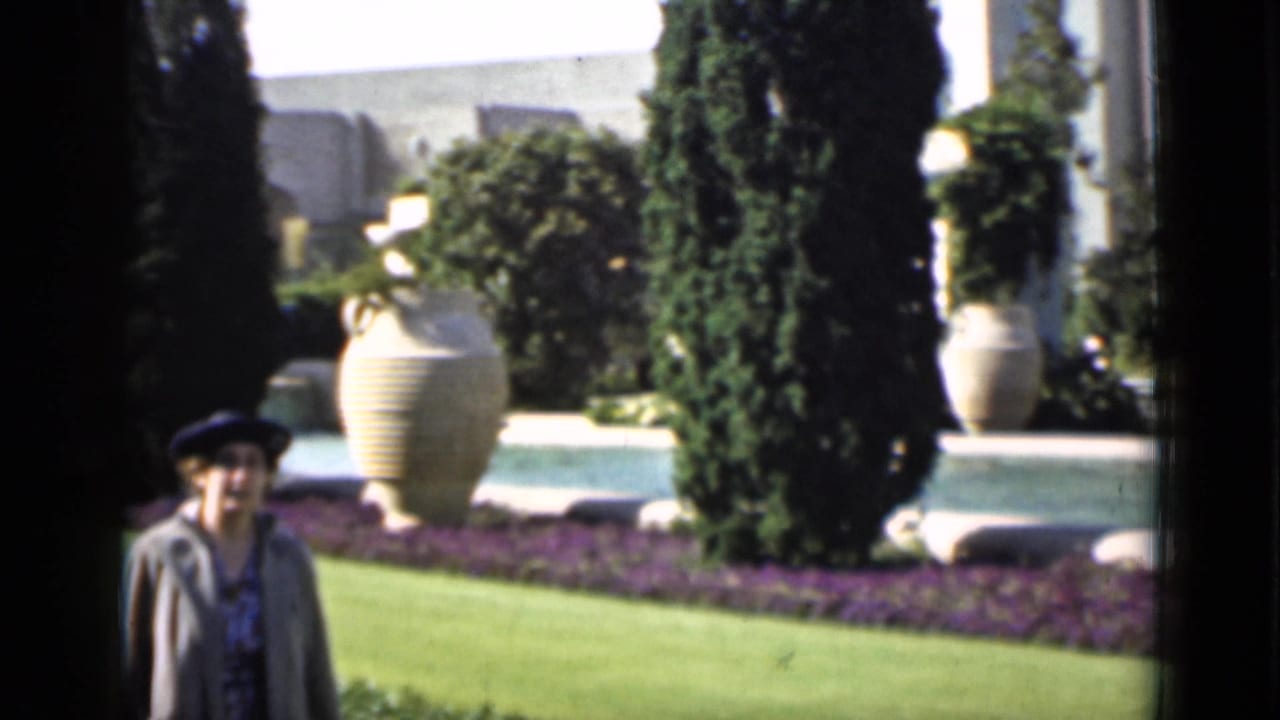 1937 San Francisco home-movie frame of a woman facing the camera in formal grounds with urns, trees, lawn, and white architecture.