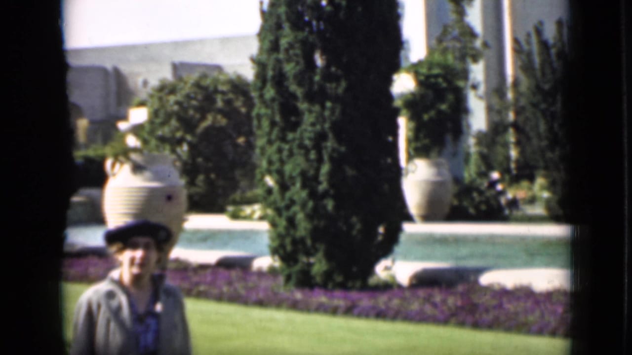 Woman in a dark hat walking through a formal garden with lawn, trees, and urn planters.