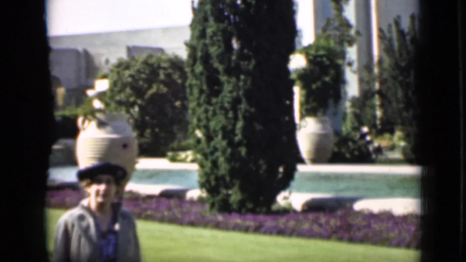 In 1937 San Francisco home-movie footage, a woman walks through a sunlit formal garden with cypress-like trees and large urns.
