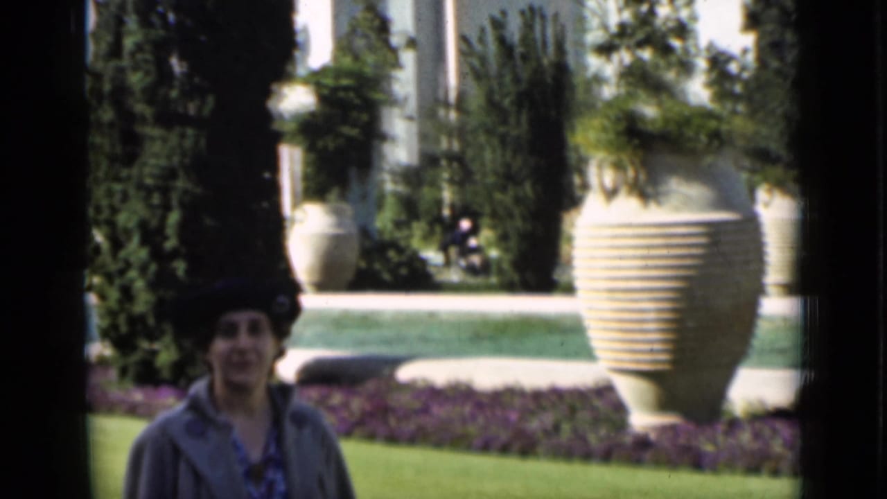 A woman appears in a formal San Francisco garden scene with a reflective pool, trimmed lawn, tall plantings, and oversized urns.