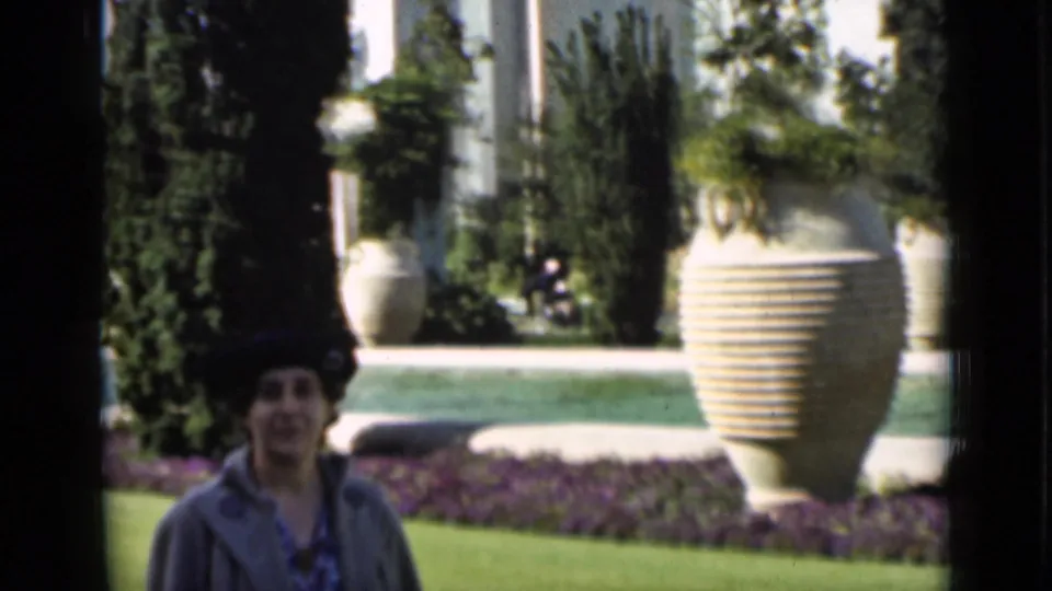 A woman appears in a formal San Francisco garden scene with a reflective pool, trimmed lawn, tall plantings, and oversized urns.