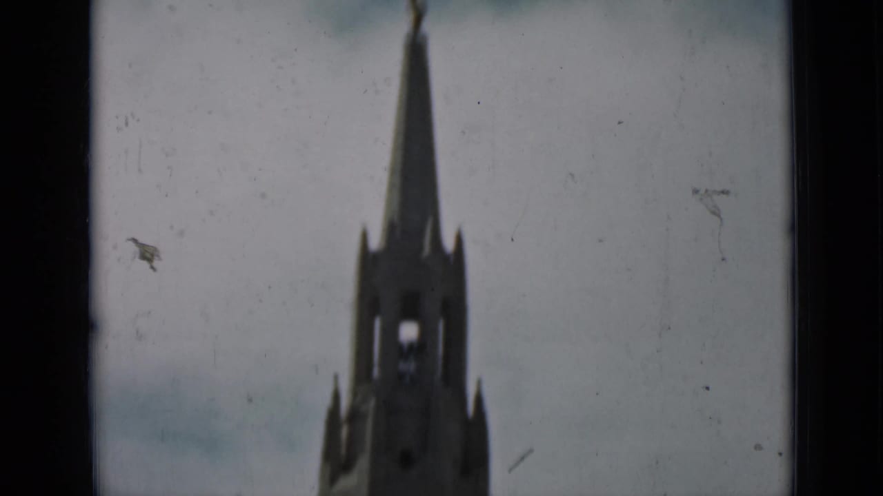 A soft-focus 8mm view shows a tall Gothic-style church or cathedral spire against a pale cloudy sky.
