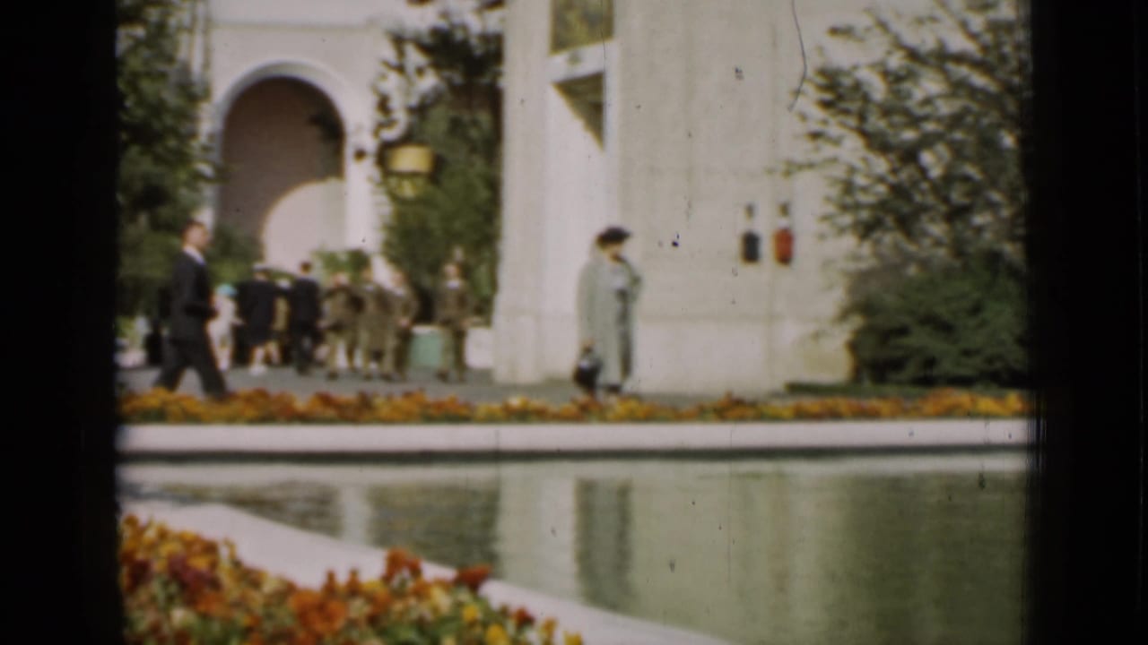 People near a reflecting pool, flower bed, shrubs, and a white arched building.