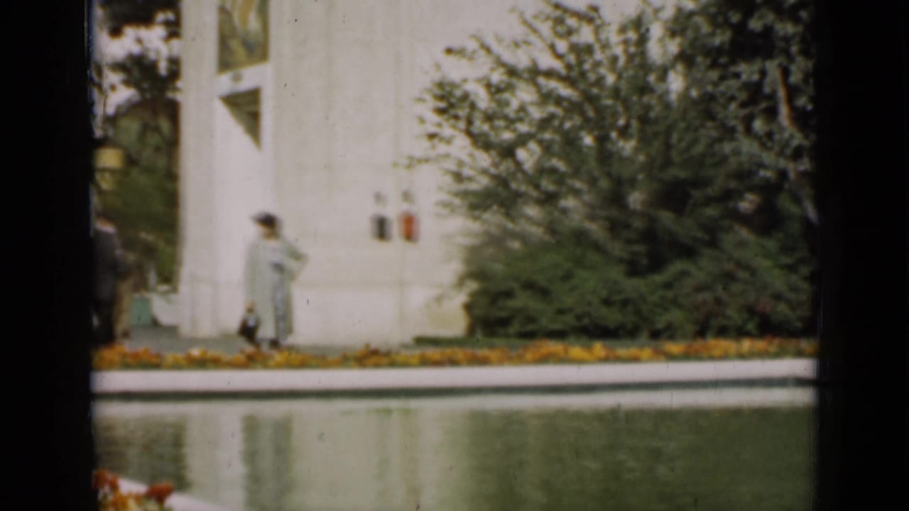A woman in a light coat and hat walks beside a reflecting pool in a formal San Francisco garden, with shrubs and a white building behind her.