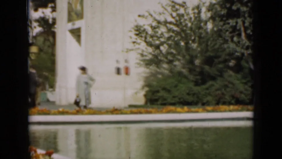 A woman in a light coat and hat walks beside a reflecting pool in a formal San Francisco garden, with shrubs and a white building behind her.