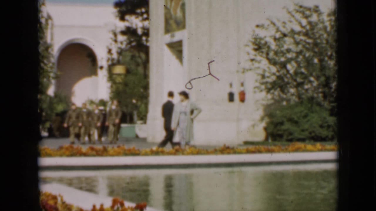 Uniformed men gather near a white arched building while two pedestrians pass beside the reflecting pool in this 1937 San Francisco garden scene.