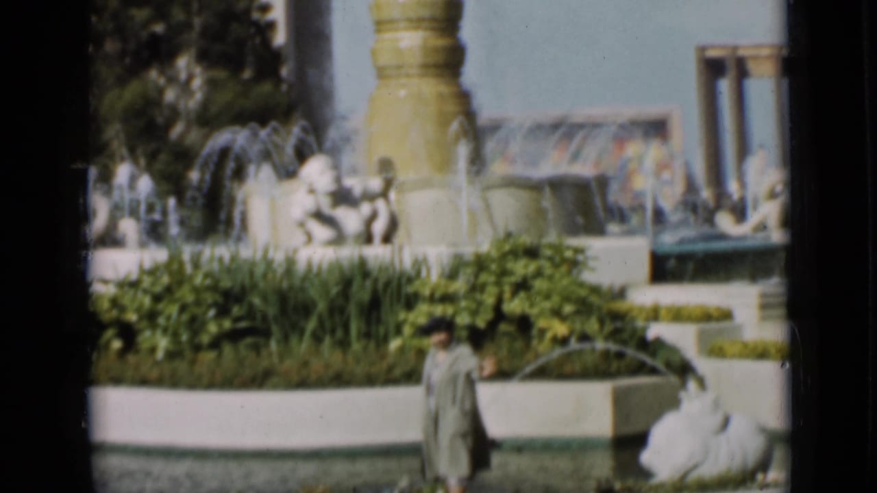 A fountain with sculpted figures and water jets rises behind planting beds while a woman stands near the pool in a San Francisco garden frame.