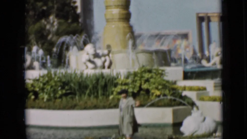 A fountain with sculpted figures and water jets rises behind planting beds while a woman stands near the pool in a San Francisco garden frame.