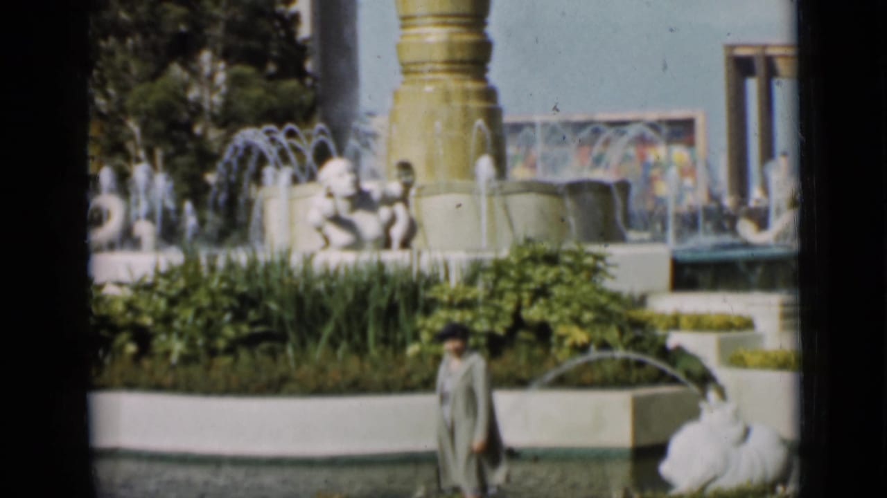Man in a light coat walking beside ornate fountains and planted beds.