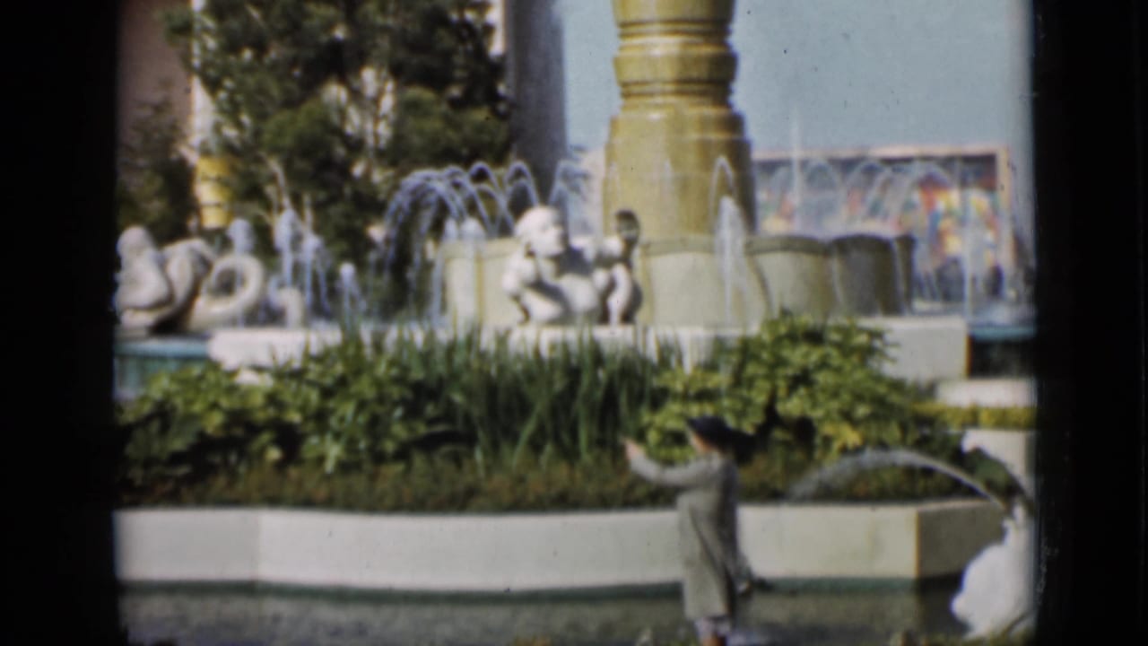 1937 San Francisco frame with a man in profile passing a formal fountain garden, white sculpture, planted beds, and a tall column.