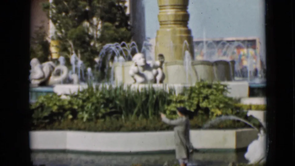 1937 San Francisco frame with a man in profile passing a formal fountain garden, white sculpture, planted beds, and a tall column.