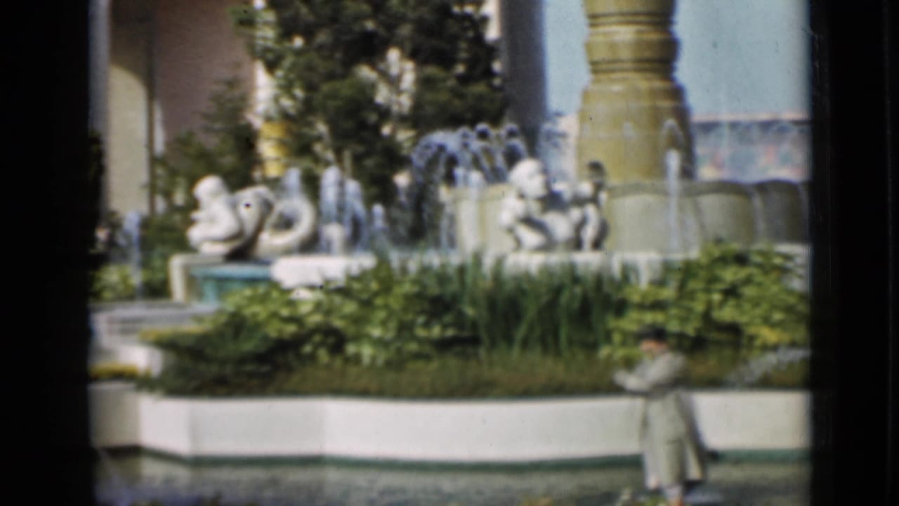 1937 San Francisco home-movie view of ornate fountain sculptures, water sprays, dense planting, and a partially visible man near the foreground.