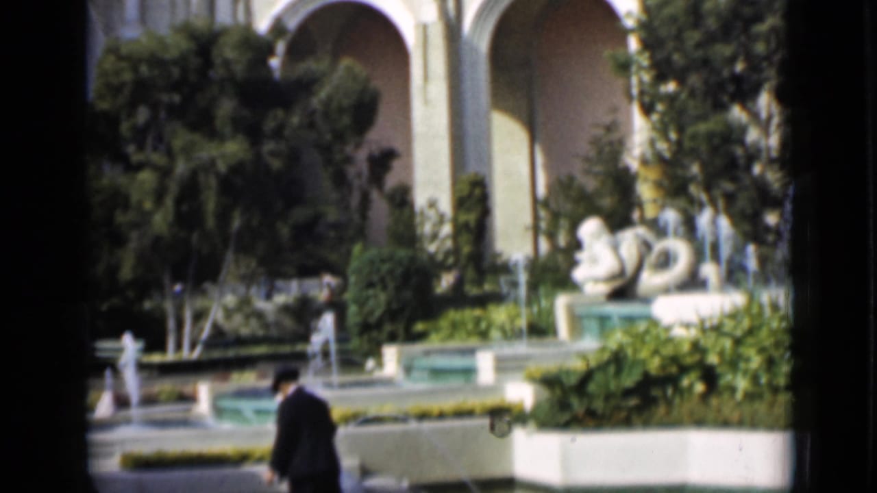 1937 San Francisco frame showing a man walking near pools, fountain sprays, white sculpture, plantings, and arched architecture in the background.