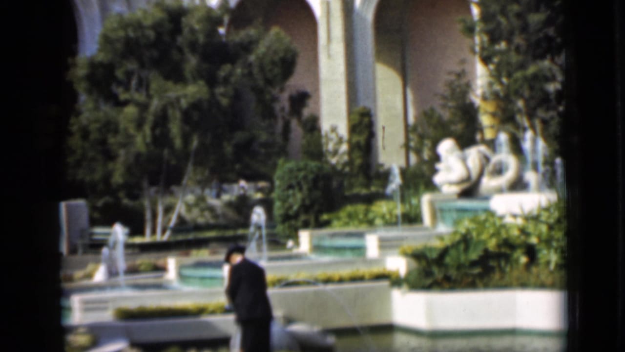 Formal garden with pools, fountains, statues, arches, and a man walking in a dark suit.