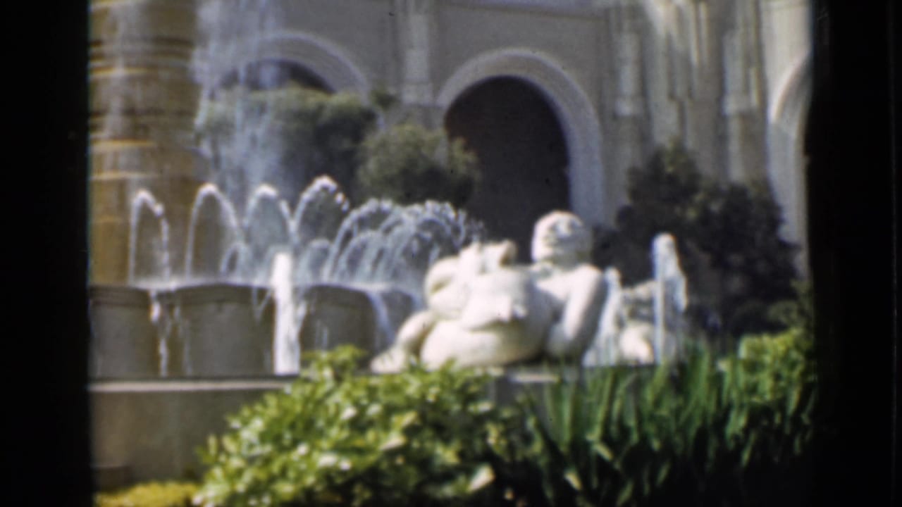 Tiered fountain with white sculpture, arched building, shrubs, and sunlit garden beds.