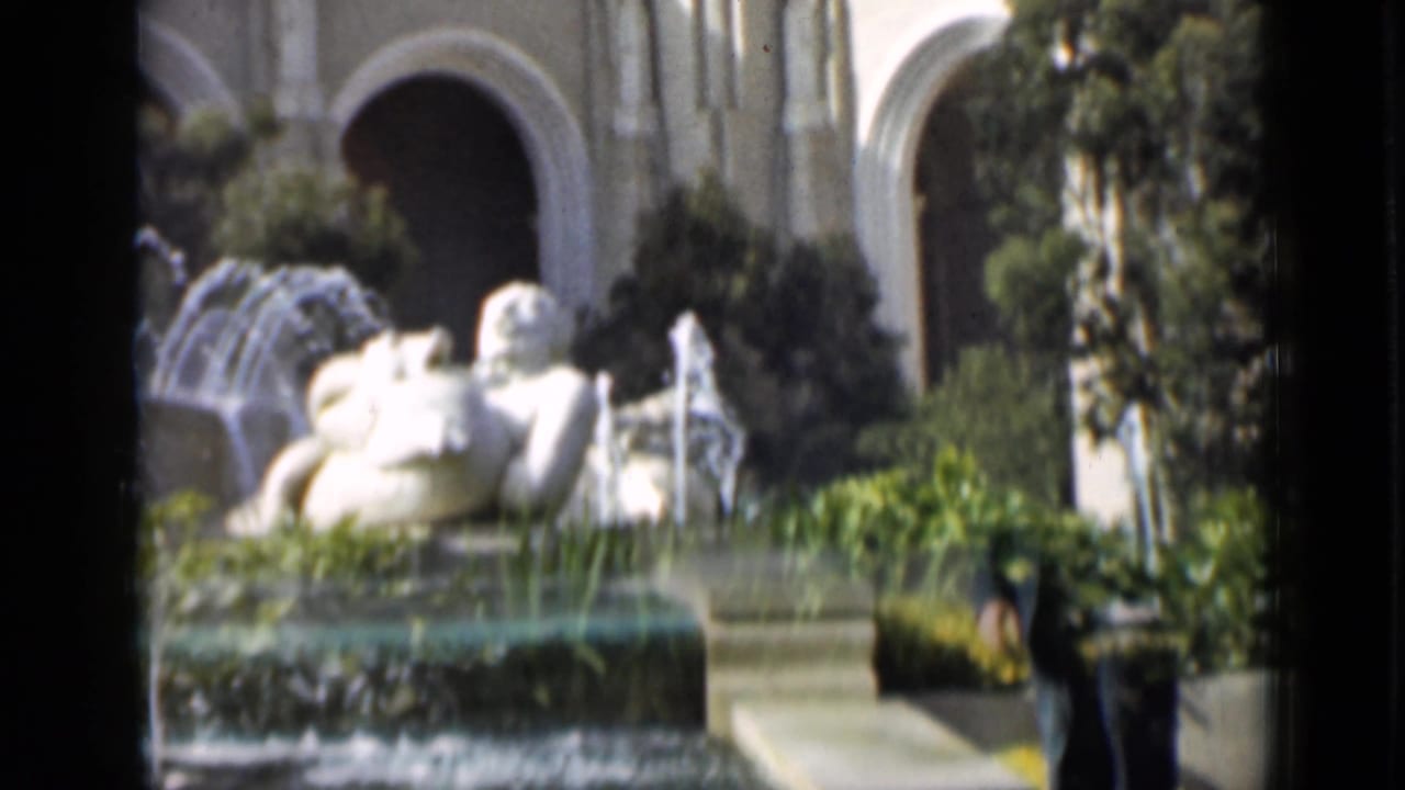 Fountain and white sculpture with arched background, shrubs, and a man entering at right.