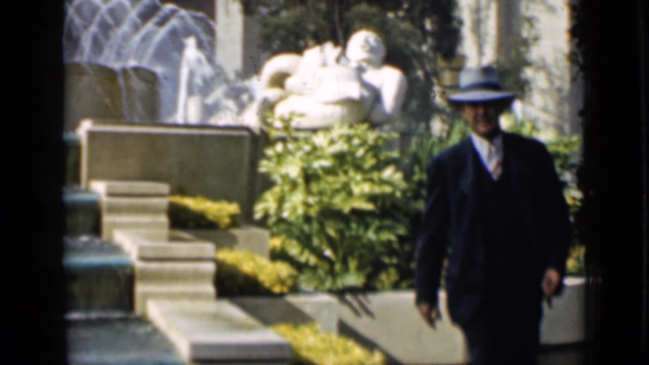 A suited man in a fedora is framed against a 1937 San Francisco garden fountain, sculpture, shrubs, and arched architecture.