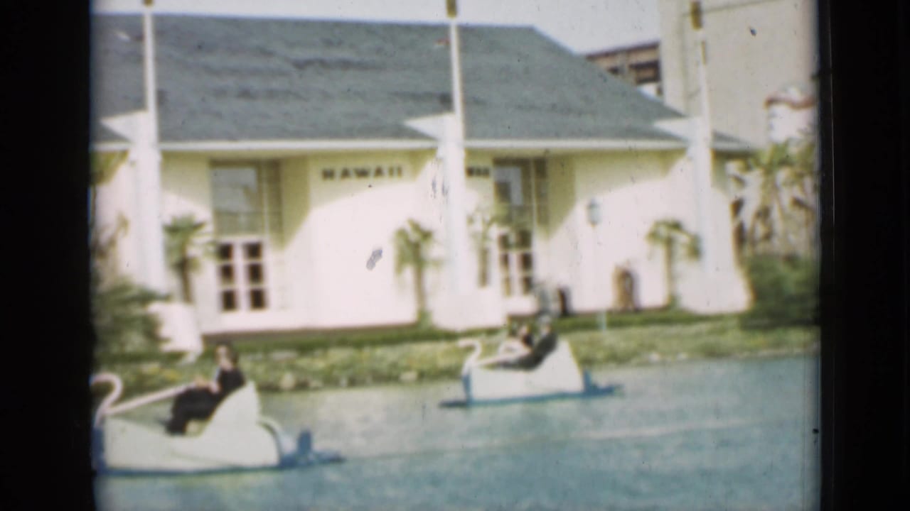 Two swan-shaped paddle boats pass a white building marked HAWAII beside a landscaped lake.
