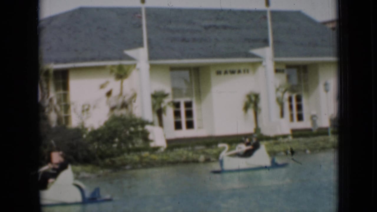 In this 1937 San Francisco frame, a swan boat passes in front of a landscaped bank and the white building marked HAWAII.