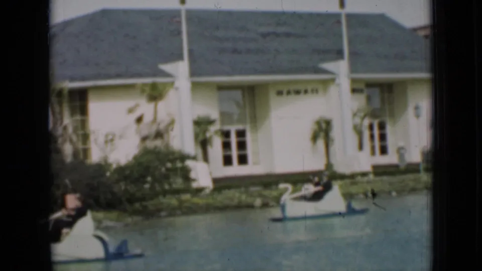 In this 1937 San Francisco frame, a swan boat passes in front of a landscaped bank and the white building marked HAWAII.