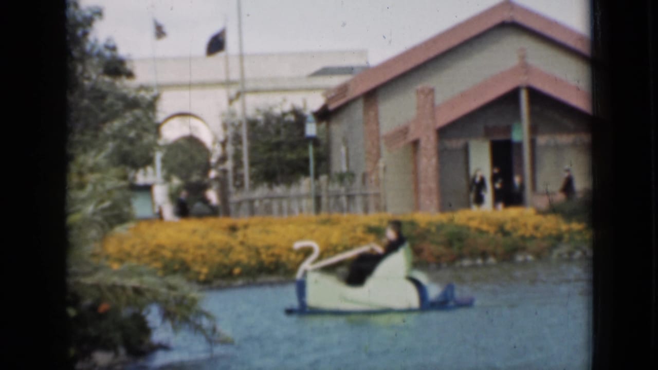 Swan-shaped paddle boat on a pond with yellow flowers and a red-roofed building behind it.