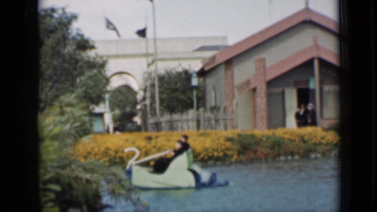 Swan-shaped paddle boat near yellow flowers with people by a small building in the background.