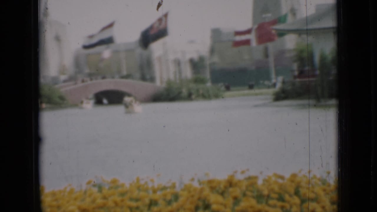 1937 San Francisco frame with a low arched bridge over the lagoon, flags above the walkway, shrubs, flowers, and a pale building.
