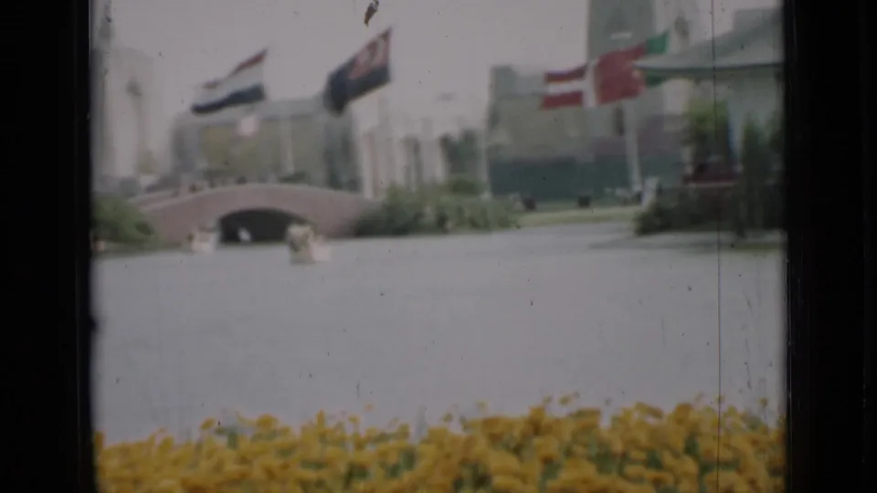 1937 San Francisco frame with a low arched bridge over the lagoon, flags above the walkway, shrubs, flowers, and a pale building.