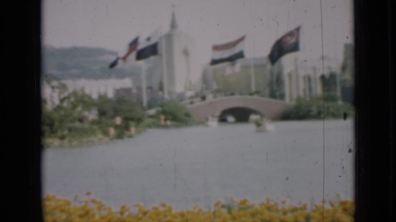 1937 San Francisco home-movie view of flags, a white building, a small arched bridge, yellow flowers, water, and distant hills.