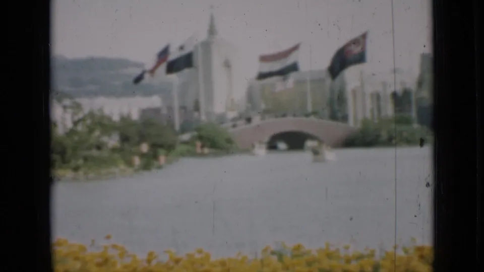1937 San Francisco home-movie view of flags, a white building, a small arched bridge, yellow flowers, water, and distant hills.