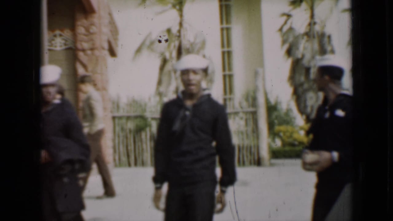 Sailors in dark uniforms and white caps walk near a building with palms and stonework.