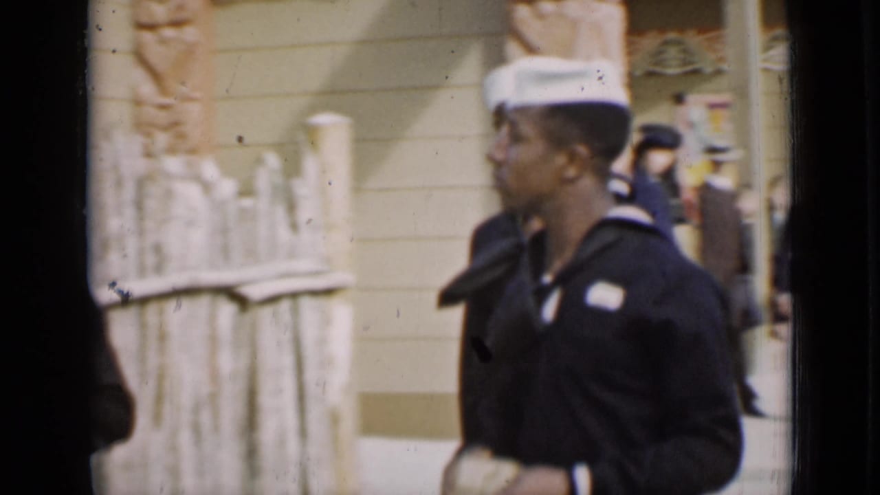 A sailor in profile passes close to the camera, with a pale fence and background pedestrians in the San Francisco 1937 footage.
