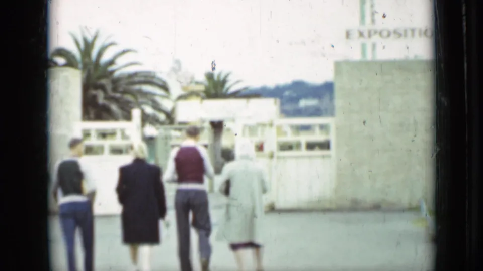 Four visitors walk away from the camera toward the Exposition Entrance in San Francisco, with palm trees and pale entrance structures visible.