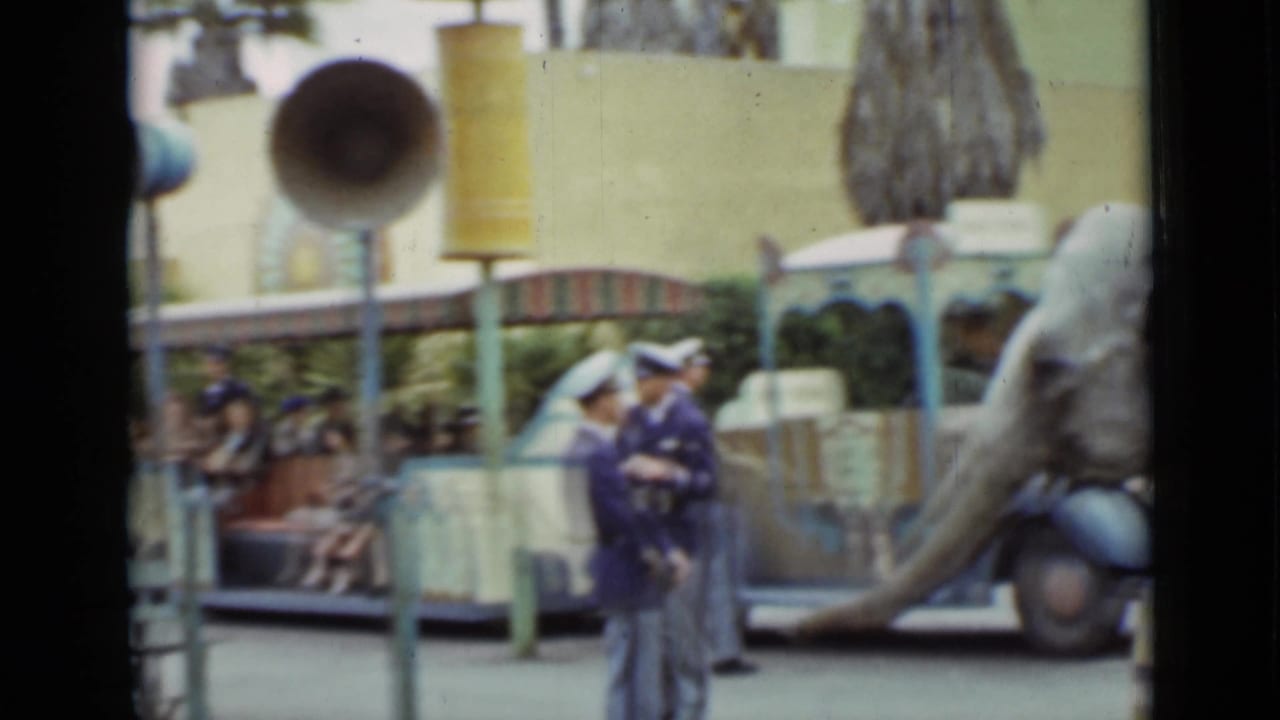 Uniformed attendants stand near a ride vehicle, benches, loudspeakers, and park guests.
