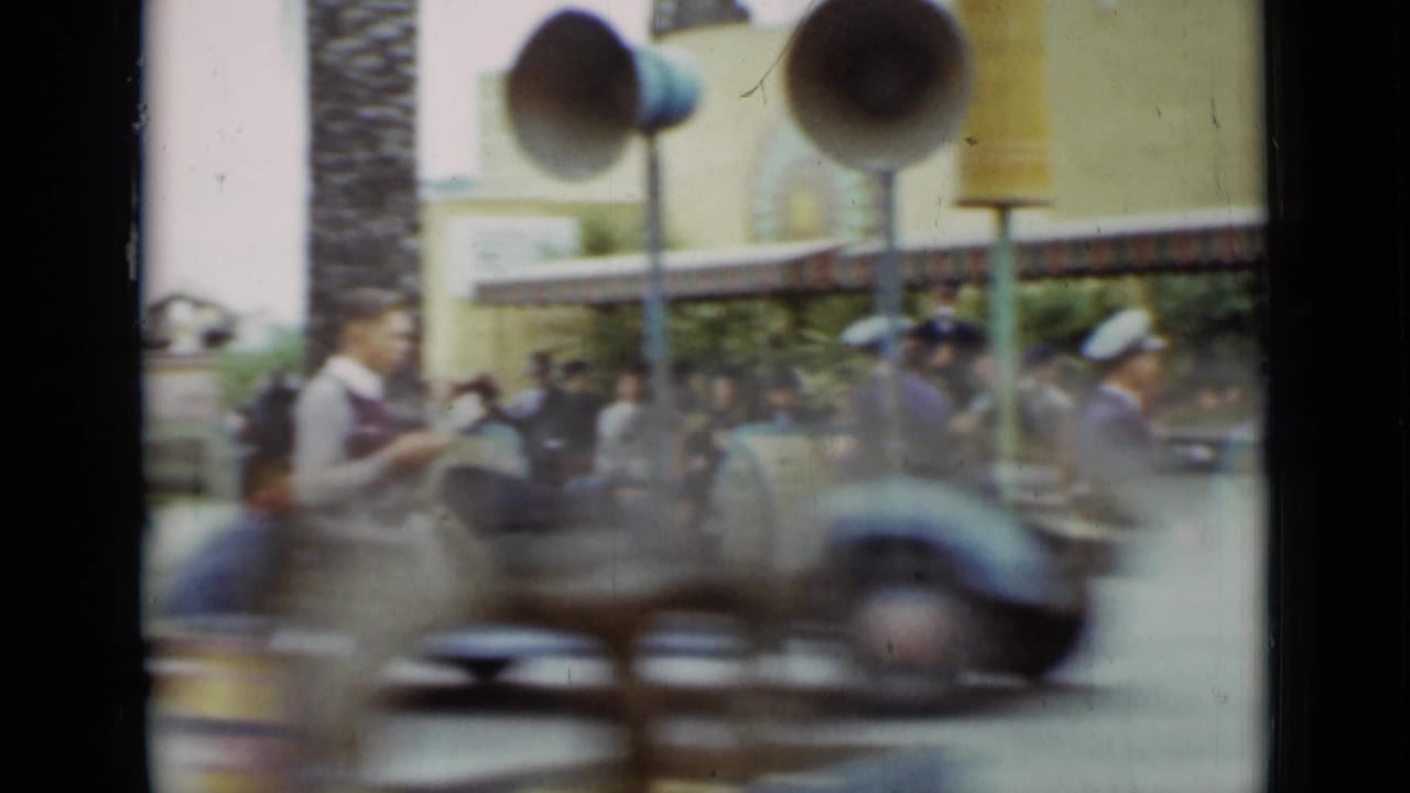 1937 San Francisco home-movie frame with motion-blurred ride vehicle crossing in front of attendants, guests, benches, and loudspeakers.