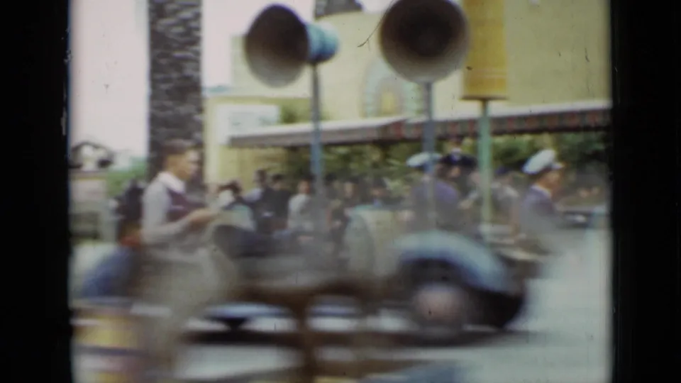 1937 San Francisco home-movie frame with motion-blurred ride vehicle crossing in front of attendants, guests, benches, and loudspeakers.