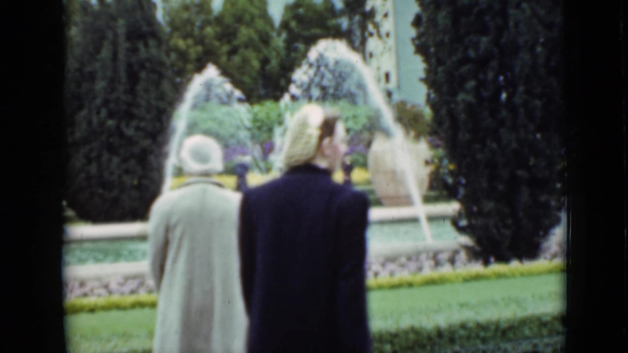 Two women in coats stand or walk near arcing fountain jets in a formal garden, supported by the San Francisco 1937 clip metadata.