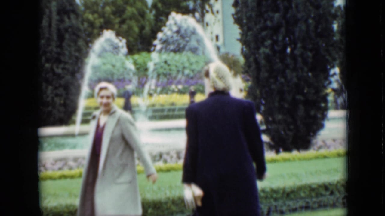 A woman in a light coat and hat smiles toward the camera as another woman in a dark coat walks near a garden fountain.