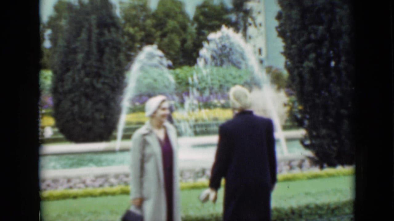 Two women in period coats pass a large fountain with arcing water, hedges, flower beds, and tall trees visible behind them.