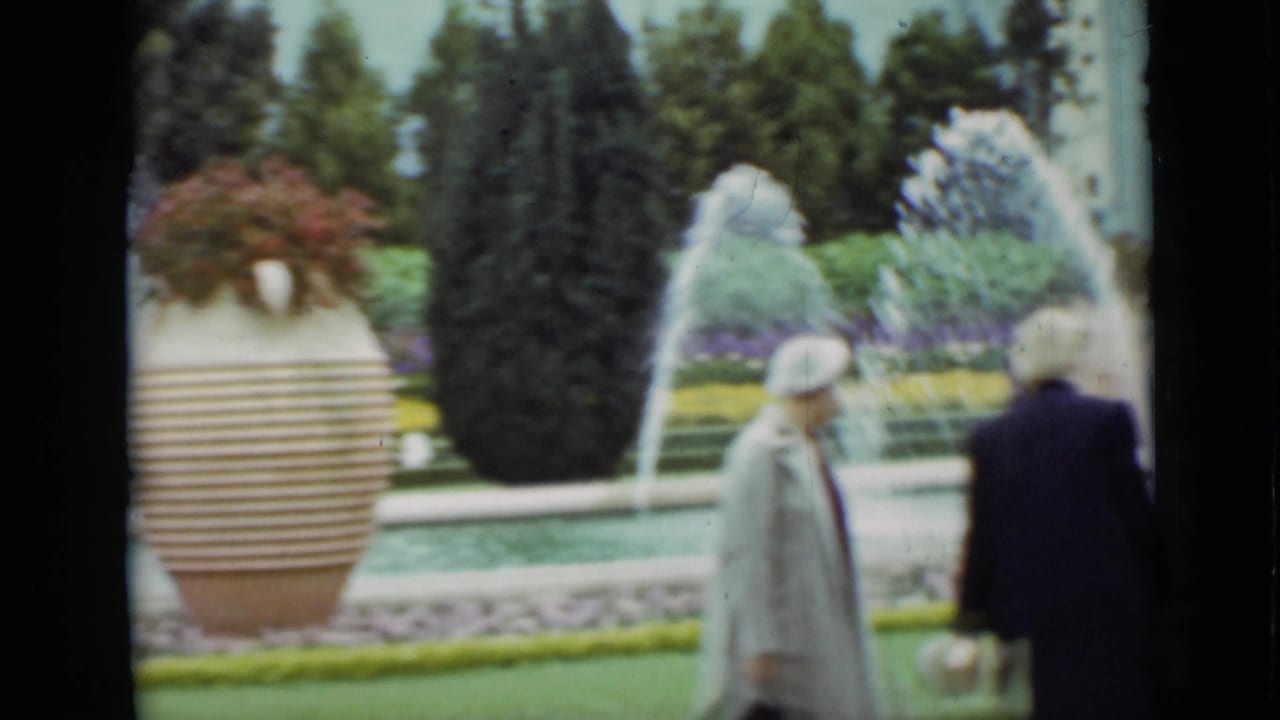 In supported 1937 San Francisco footage, two adult women in coats and hats walk by arcing fountain jets, lawns, hedges, and a striped planter.