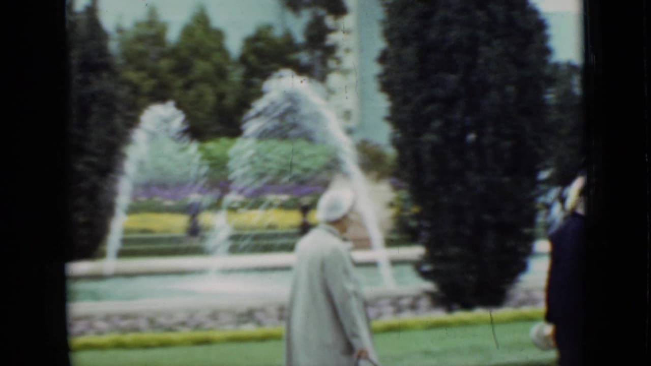 A woman in a light coat and hat walks through the garden in the 1937 San Francisco home-movie scene, with fountain jets and clipped plantings behind her.
