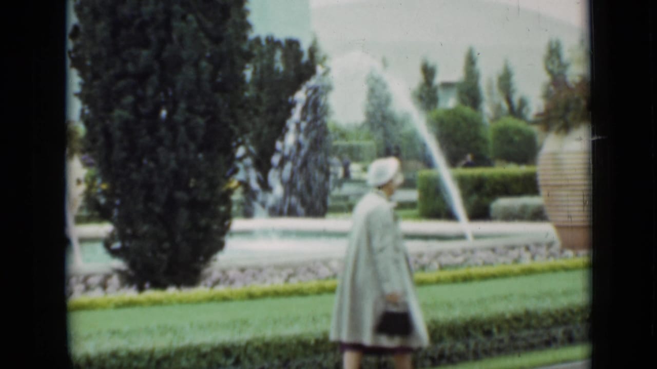 A woman in a light coat and hat crosses the garden frame, with arcing water jets, hedges, trees, and a striped planter visible in the 1937 San Francisco footage.