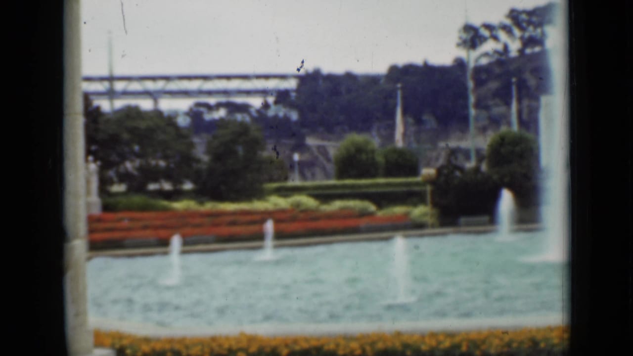 Formal fountain basin with small jets, flower beds, shrubs, hillside, and distant bridge-like structure.