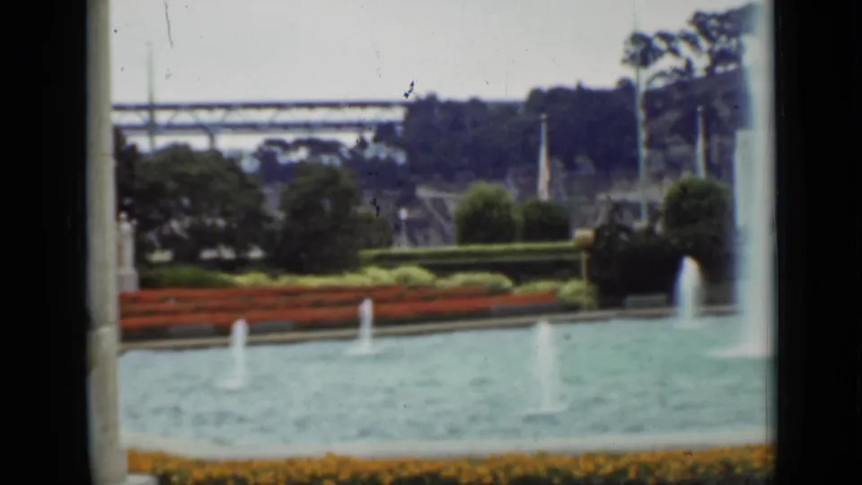 1937 San Francisco park view with fountain water, red flower beds, clipped hedges, trees, and a distant bridge-like structure beyond the garden.