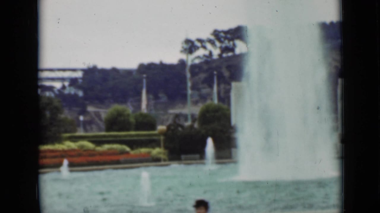 Large fountain plume beside smaller jets, garden plantings, trees, flags, and a partial passerby.