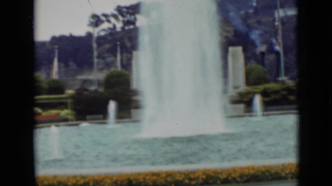 San Francisco park fountain in 1937, showing a central water plume, smaller jets, clipped shrubs, flower border, trees, and a nearby flagpole.